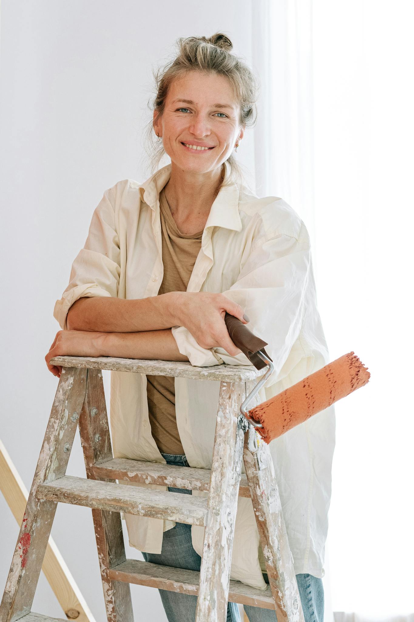 Adult woman in casual attire stands confidently on ladder with paint roller for DIY home renovation.